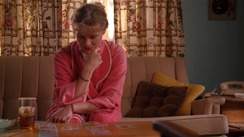 A scene from Mad Men. Anna Draper, a blonde white woman in her thirties, sits on a couch in a pink bathrobe. She rests her chin in her hand as she looks down at tarot cards laid out on a coffee table.