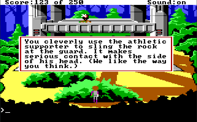 Roger crouches behind a bush facing a metallic tower structure. A guard stands on the structure, being hit in the face with something. Game text: "You cleverly use the athletic supporter to sling the rock at the guard. It makes serious contact with the side of his head. (We like the way you think.)"